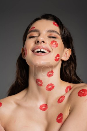 Foto de Excited woman with bare shoulders and red lipstick marks on body and face laughing with closed eyes isolated on grey - Imagen libre de derechos