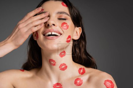 Foto de Cheerful woman with red lip prints on face and bare shoulders covering eye with hand isolated on grey - Imagen libre de derechos