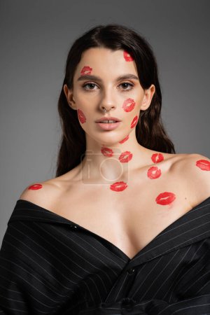 Foto de Young brunette woman with lip prints on face and bare shoulders looking at camera isolated on grey - Imagen libre de derechos