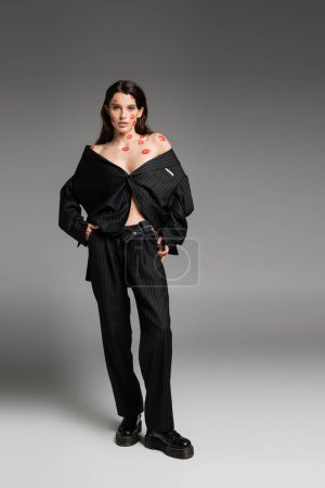 Photo for Full length of brunette woman with red kiss prints wearing black oversize suit and looking at camera on grey - Royalty Free Image