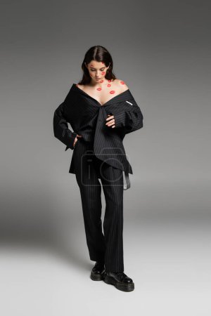 Photo for Full length of woman with red lip prints on bare shoulders and face standing in black oversize suit and rough boots on grey - Royalty Free Image