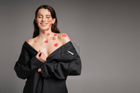 Foto de Seductive woman with red kiss prints on face and naked shoulders posing in oversize blazer isolated on grey - Imagen libre de derechos