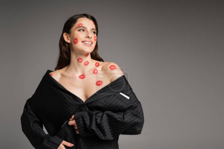 Foto de Cheerful young woman with red kisses on face and body looking away while posing in oversize blazer isolated on grey - Imagen libre de derechos