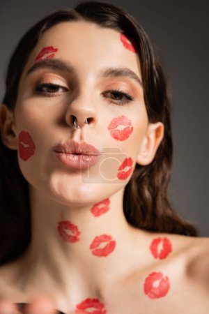 portrait of pretty woman with makeup and red kiss prints pouting lips while looking at camera isolated on grey
