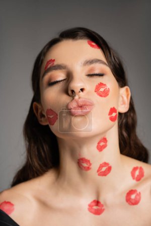 portrait of sexy woman with closed eyes and red kiss prints on face pouting lips isolated on grey