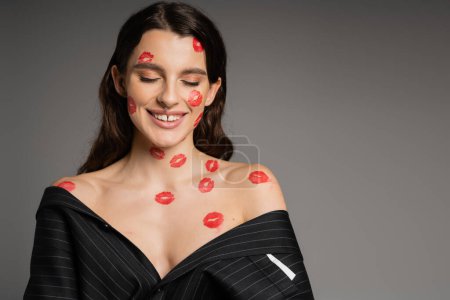 Foto de Happy young woman with red lipstick marks on face and bare shoulders smiling with closed eyes isolated on grey - Imagen libre de derechos