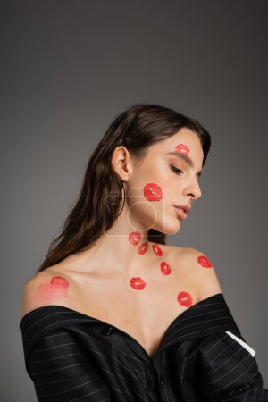 portrait of brunette woman with red kiss prints on naked shoulders and face isolated on grey