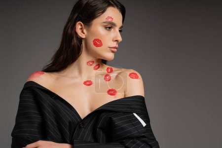 Foto de Brunette woman with red kiss prints posing in black oversize jacket with bare shoulders isolated on grey - Imagen libre de derechos