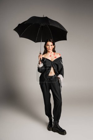 full length of sexy woman with red kiss prints wearing oversize suit while holding umbrella on grey background