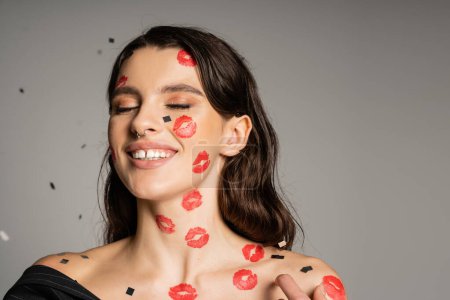 young brunette woman with lip prints on face and body smiling with closed eyes on grey background