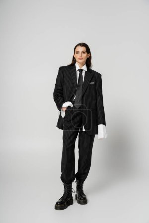 Photo for Full length of brunette woman in stylish formal wear and rough leather boots posing with hand in pocket on grey - Royalty Free Image