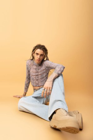 Téléchargez les photos : Stylish queer person in jeans and crop top sitting on yellow background - en image libre de droit
