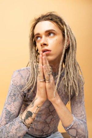 Tattooed queer person doing praying hands gesture on yellow background 