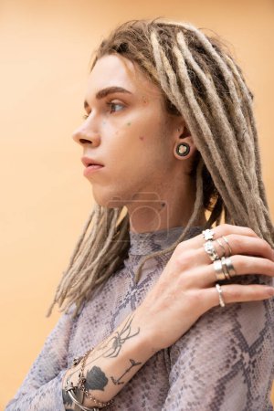 Portrait of tattooed queer person with dreadlocks touching shoulder isolated on yellow 