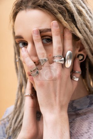 Nonbinary person with silver rings on fingers touching face isolated on yellow