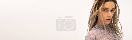 Young tattooed queer person with hairstyle looking at camera isolated on white, banner 