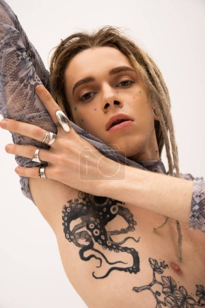 Portrait of tattooed queer person in accessories touching arm isolated on white 