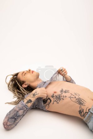 Tattooed nonbinary person touching crop top with animal print while lying on white background