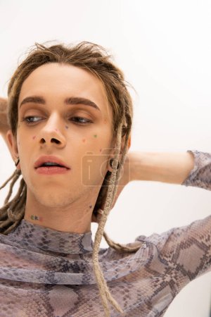 Portrait of tattooed queer person touching dreadlocks and looking away isolated on white 