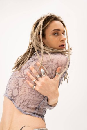 trendy queer model in silver rings and snakeskin print top looking at camera isolated on white