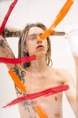 young tattooed queer person painting on transparent surface with paintbrush on white background