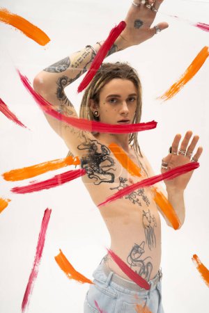 queer person with tattooed body and dreadlocks looking at camera near glass with brush strokes on white background