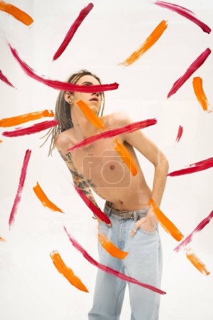 queer person with shirtless tattooed body holding hands in pockets of jeans behind glass with colorful paint strokes on white background
