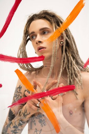 young queer person with dreadlocks and tattooed body looking away near colorful paint spills on white background