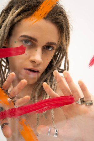 Foto de Portrait of queer model with dreadlocks and silver rings looking at camera near paint strokes on white background - Imagen libre de derechos