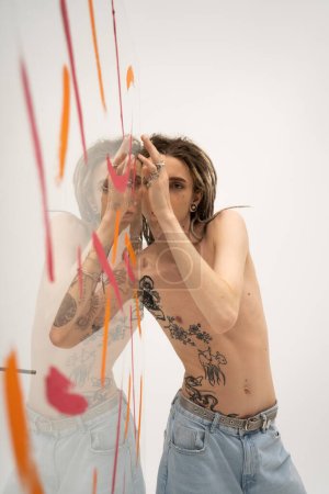 Téléchargez les photos : Shirtless tattooed queer person in jeans posing near glass with colorful paint on white background - en image libre de droit