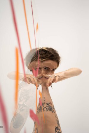 smiling tattooed queer model touching glass with paint strokes while looking at camera on white background