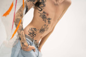 cropped view of queer model with tattooed body posing with hands in pockets of jeans near glass with paint strokes on white background t-shirt #636817136