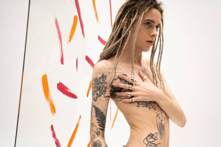 shirtless queer person with dreadlocks touching tattooed torso near multicolored paint strokes on white background magic mug #636817240