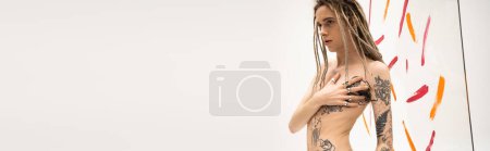 nonbinary person with dreadlocks touching shirtless tattooed body near multicolored brush spills on white background, banner