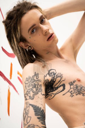 young tattooed nonbinary model with dreadlocks looking at camera near colorful paint strokes on white 