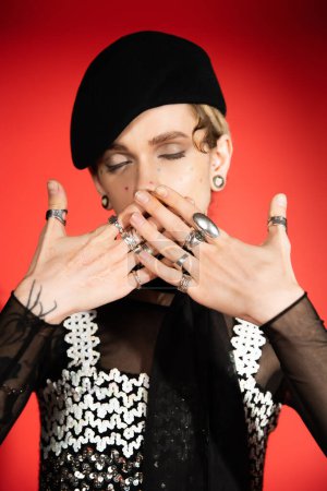 queer person with closed eyes covering mouth with hands in silver rings on red background