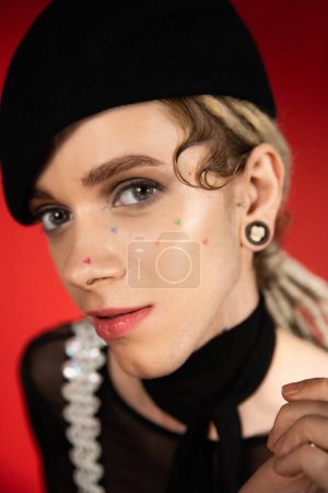 close up portrait of tattooed queer person with makeup looking at camera on red background