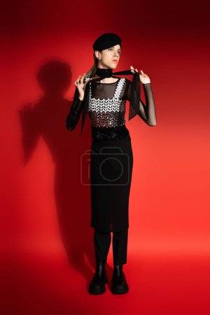 full length of nonbinary model in beret and elegant top tying neckerchief on red background with shadow