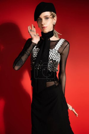 tattooed queer model in elegant attire waving hand and looking at camera on red background with shadow