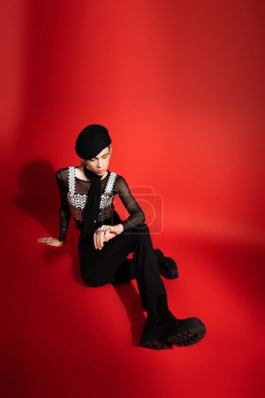 full length of young nonbinary person in black beret and top with sequins sitting on red background