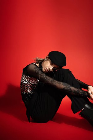 tattooed queer model in black elegant attire sitting and looking at camera on red background