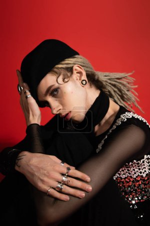 young queer person in silver finger rings and black beret looking at camera isolated on red