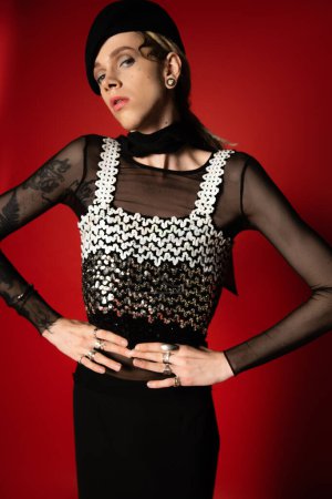 tattooed nonbinary person in elegant clothes and black beret posing with hands on waist on red background