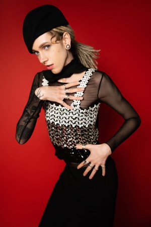 elegant nonbinary model in black beret holding hand on chest while looking at camera on red background