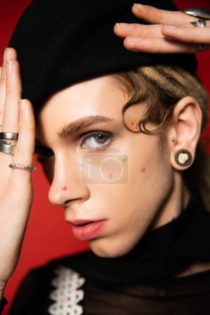 close up portrait of nonbinary model with makeup and finger rings adjusting black beret on red background