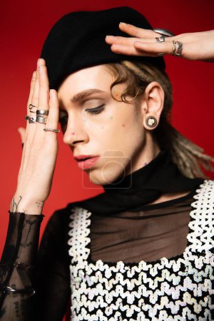 portrait of queer model in elegant top and silver rings adjusting black beret isolated on red