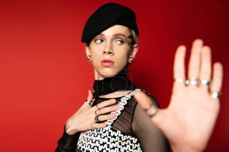 elegant queer person in black beret and silver rings holding hand on chest and showing stop gesture on red background