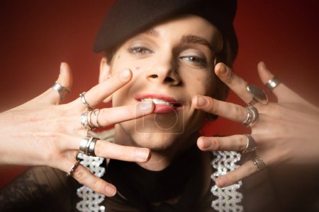 smiling queer person in black beret and silver rings posing with hands near face on dark red background