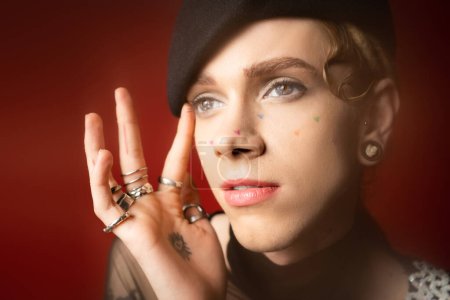 portrait of queer model in black beret and silver rings touching face and looking away on dark red background