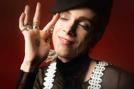 portrait of nonbinary person in stylish attire and silver rings touching face while smiling with closed eyes isolated on dark red